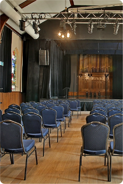 Image d'une salle de spectacle où l'on voit des chaises en alignement, une scène bordée de rideaux noirs en arrière plan et un système d'éclairage professionnel suspendu